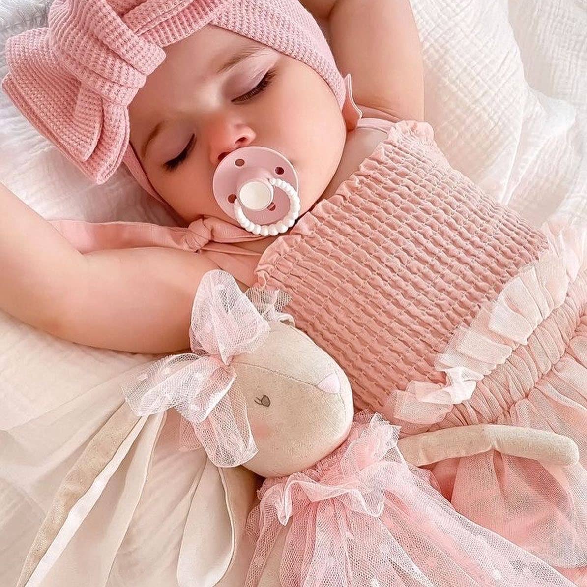 A baby girl sleeping with a pink pacifier and a soft doll in pink and white clothing