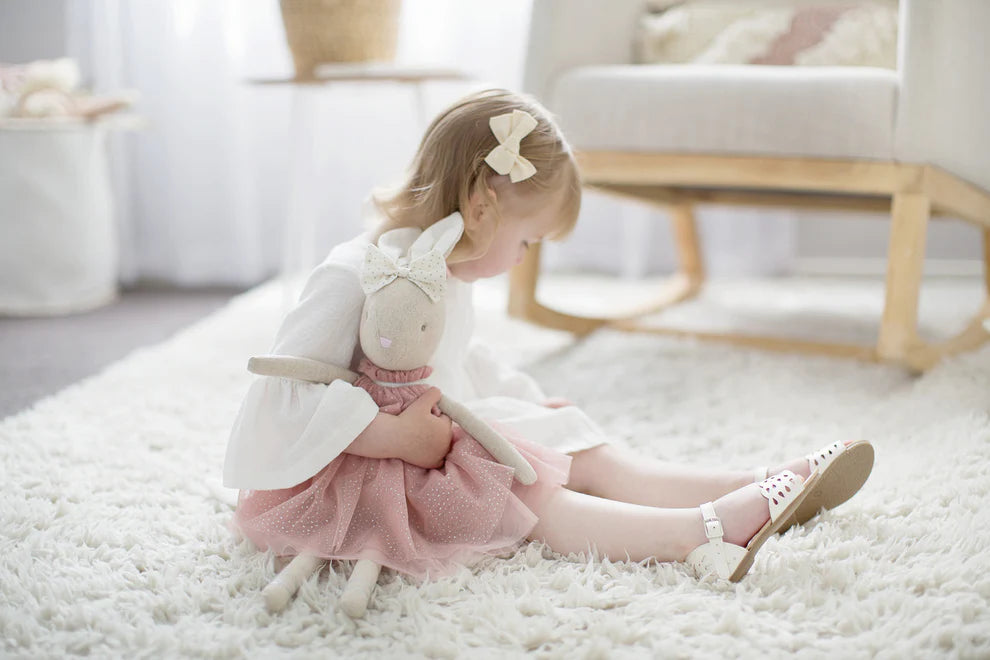 Young girl in a white dress with a pink tulle skirt, playing with a soft plush bunny toy on a carpeted floor.