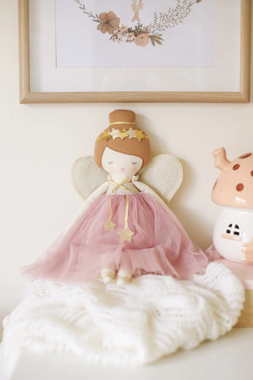 Mia Fairy Doll wearing a blush pink dress with golden wings and star accents, displayed on a cozy white blanket with soft lighting